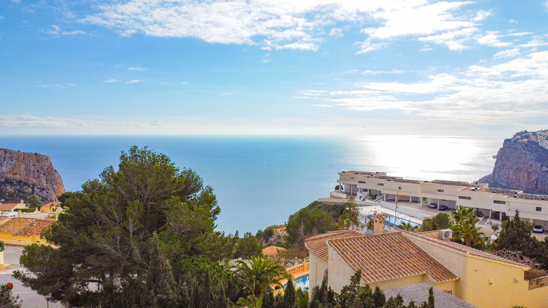 Panoramic Mediterranean Sea view from Cumbre del Sol, featuring coastal cliffs, modern and traditional architecture, and lush pine trees under a bright sky.