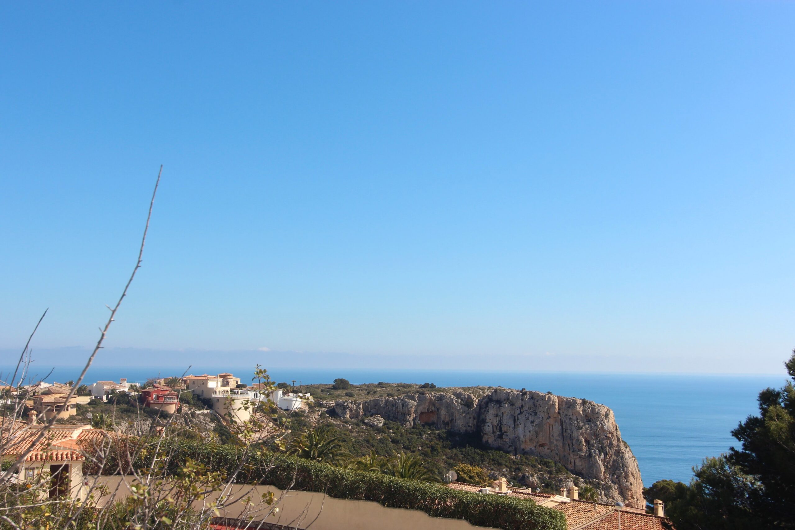Panoramic view from the property overlooking the Mediterranean Sea and rugged cliffs, featuring traditional terracotta rooftops and lush coastal vegetation under a clear blue sky.