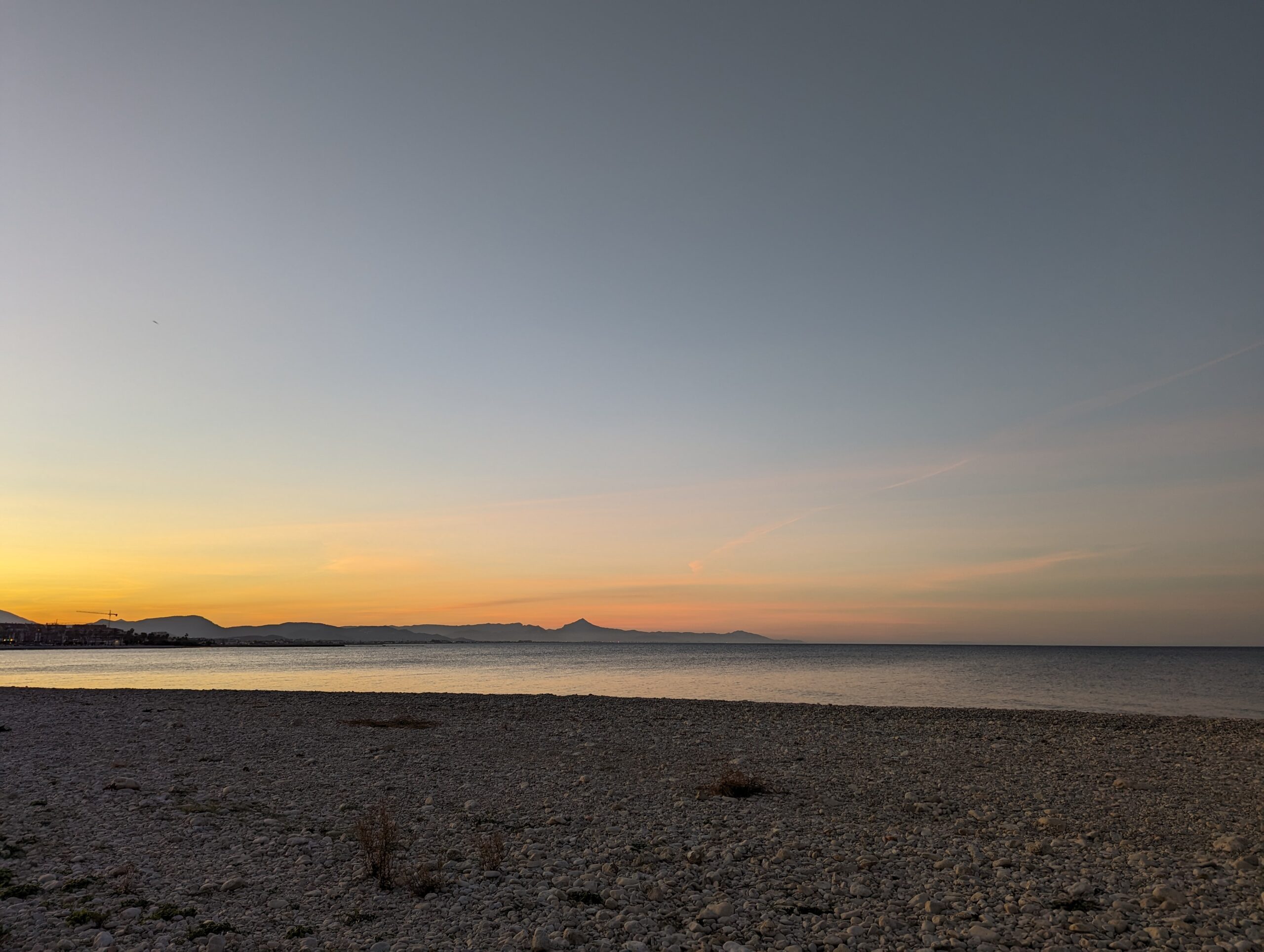 Panoramic view of a pebble beach in Denia at sunset, featuring the Mediterranean Sea and distant mountain silhouettes under a gradient sky.