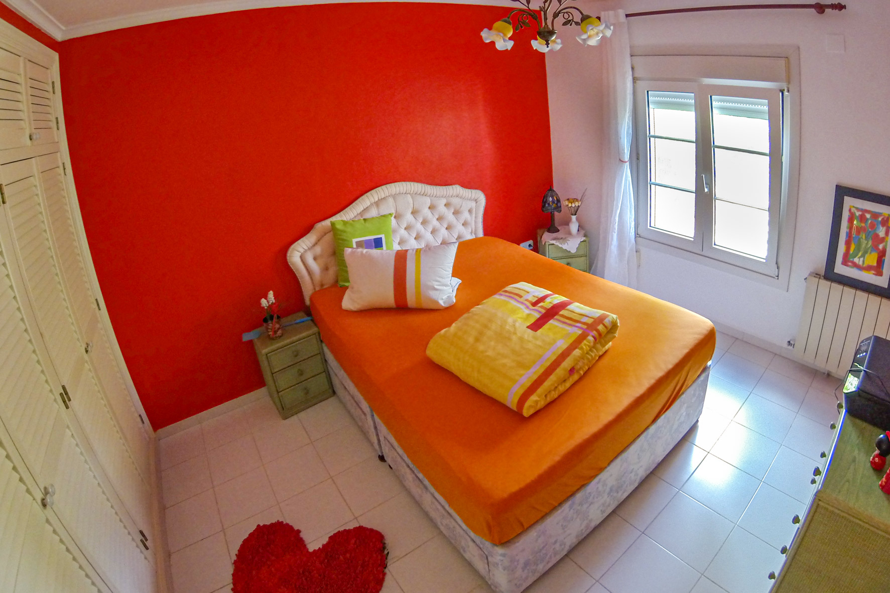 Bright bedroom featuring a red accent wall, double bed with orange linens, and upholstered headboard.