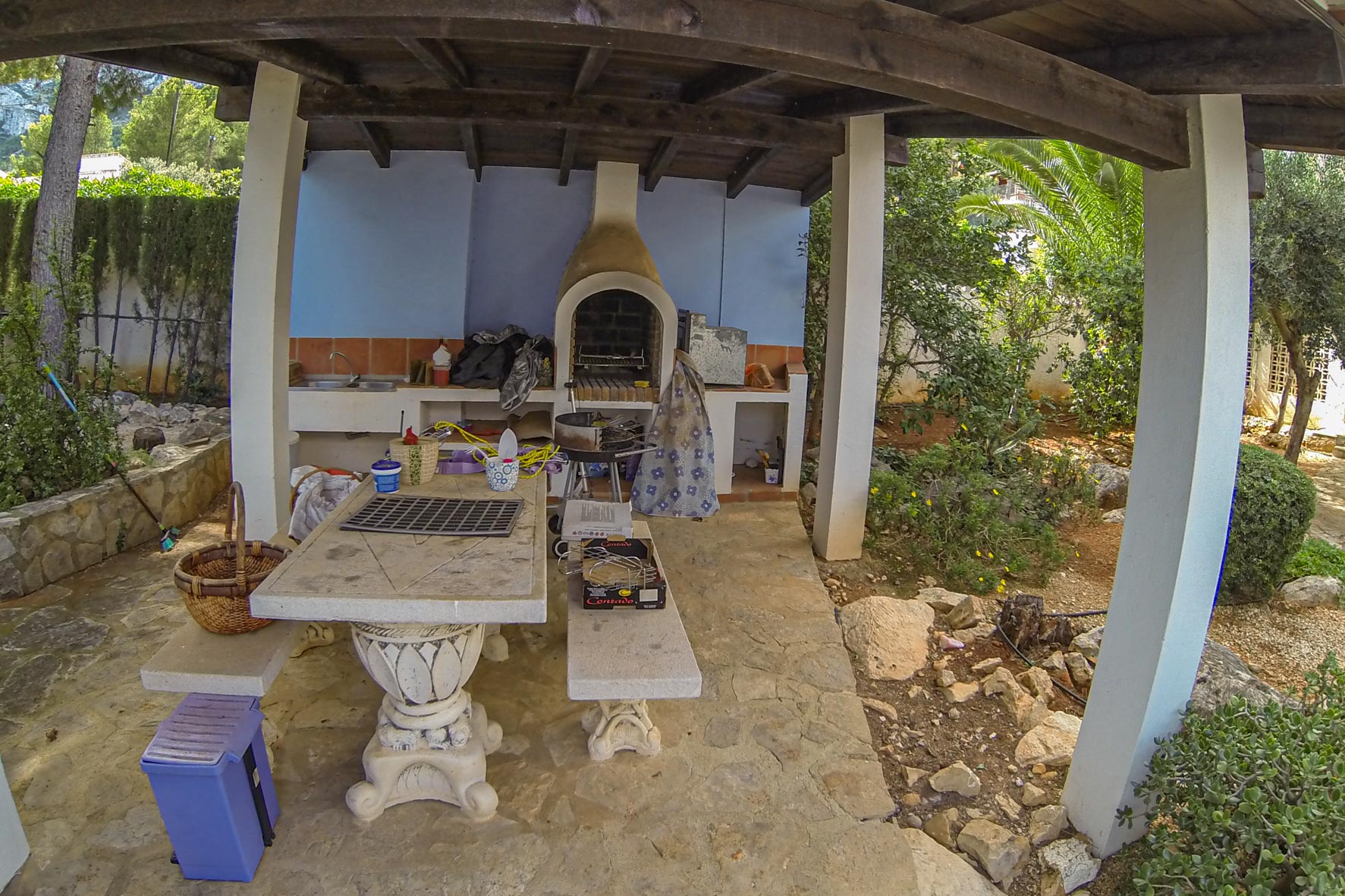 Covered barbecue area with brick oven and stone table, nestled within a lush garden.