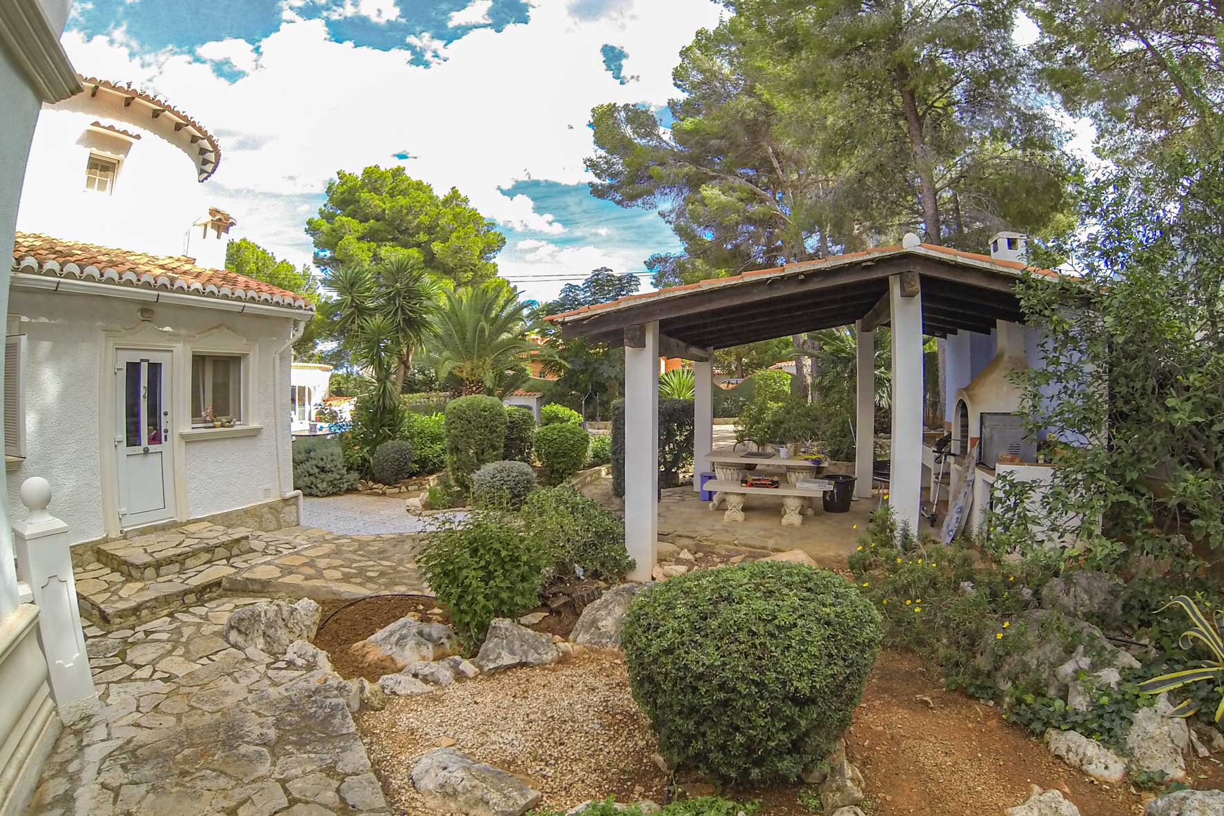 Covered barbecue area with wood-fired oven and stone table in a Mediterranean garden.