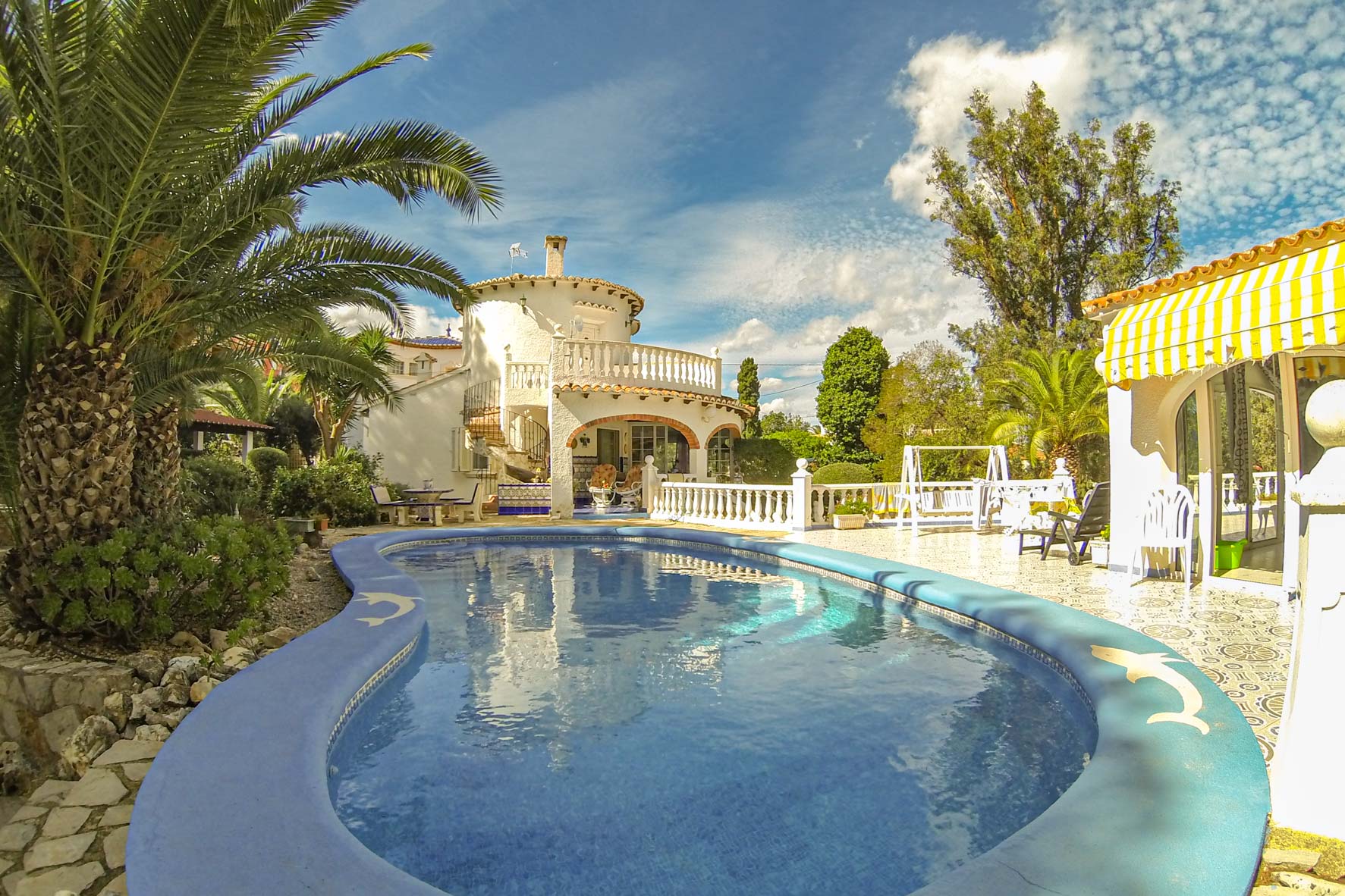 View of the turquoise blue pool with dolphin-decorated border and the facade of the two-story white villa.