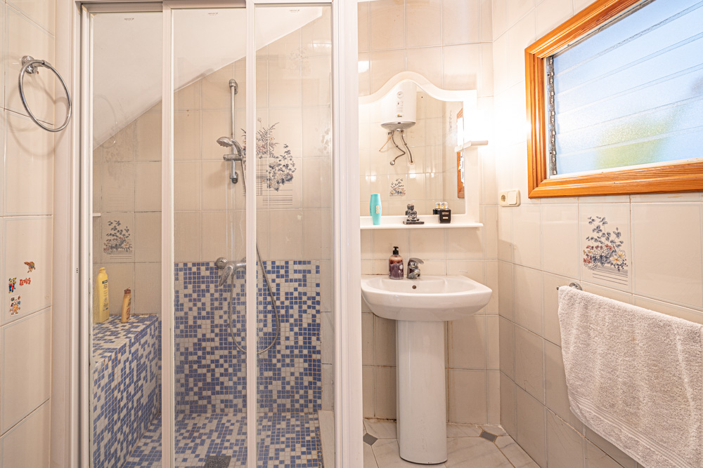 Bathroom featuring a walk-in shower with blue mosaic tiling, a classic pedestal sink, and tiled walls with decorative floral motifs and a wooden window frame.