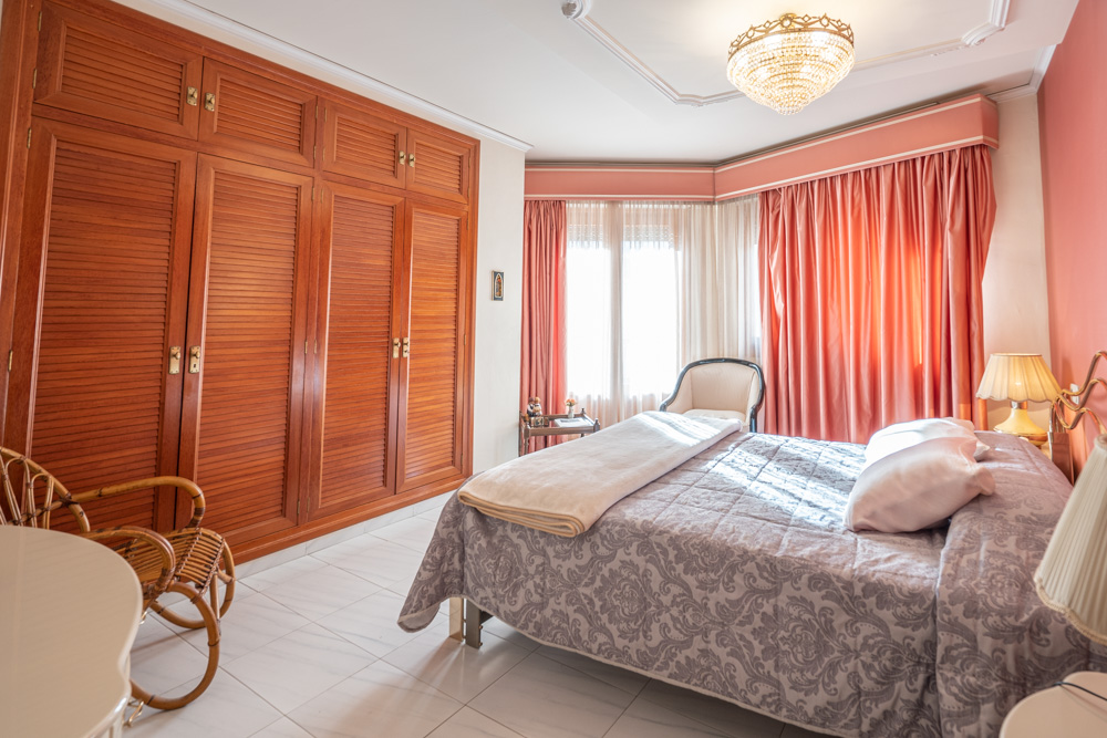 Bedroom featuring floor-to-ceiling wooden wardrobes with louvered doors, a large window with layered curtains, and a classic crystal chandelier.