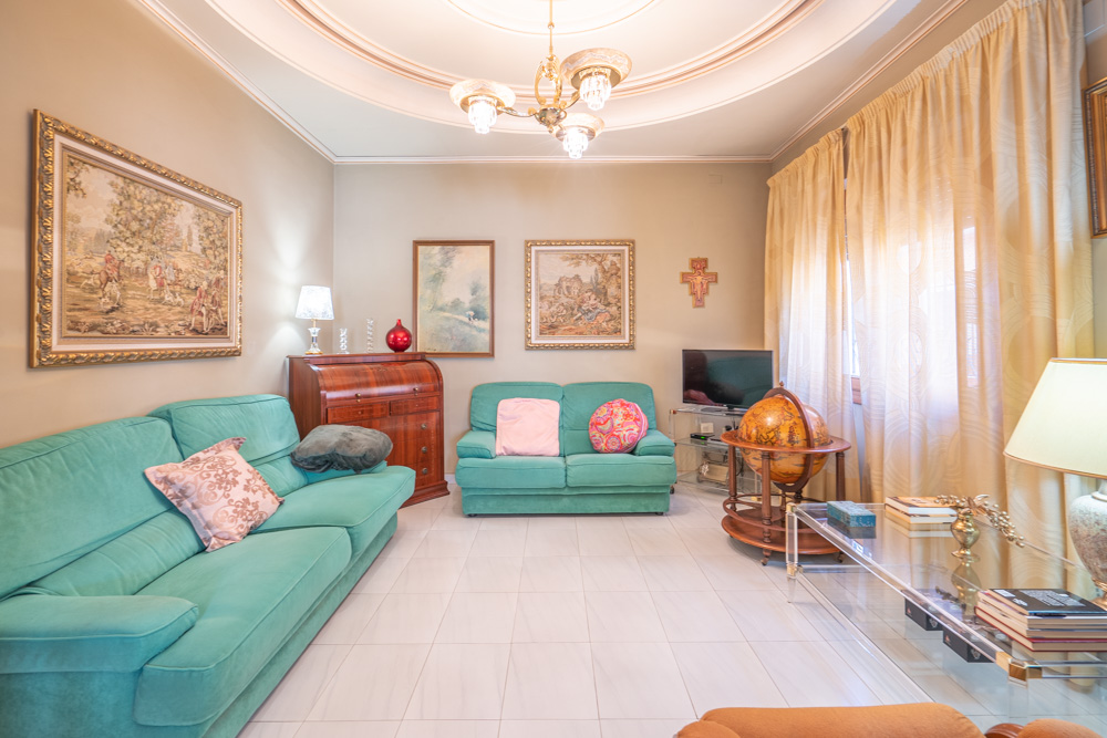 Bright living room featuring turquoise sofas, white tiled flooring, and decorative circular ceiling molding. Includes classic wooden furniture, a roll-top desk, and framed artwork.