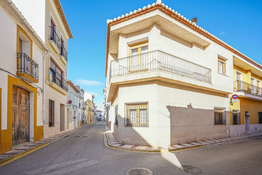 Exterior view of the corner townhouse featuring a white facade, wrap-around balcony with iron railings, and traditional windows on a quiet, sunlit village street.