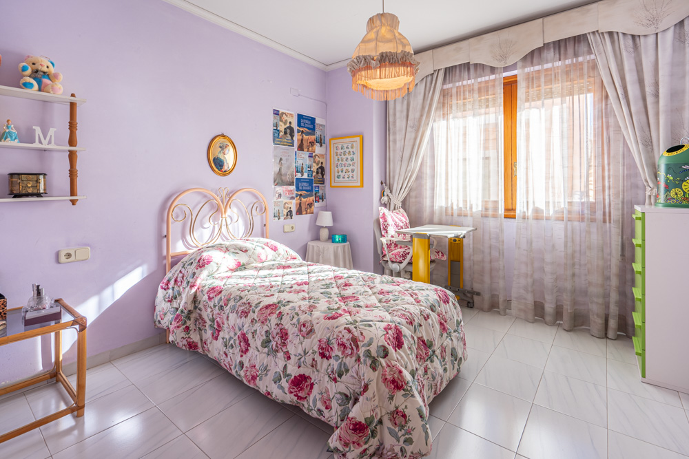 Single bedroom featuring lavender walls, light tiled flooring, and a wrought iron headboard. A study desk is positioned by the wooden window, offering natural light.