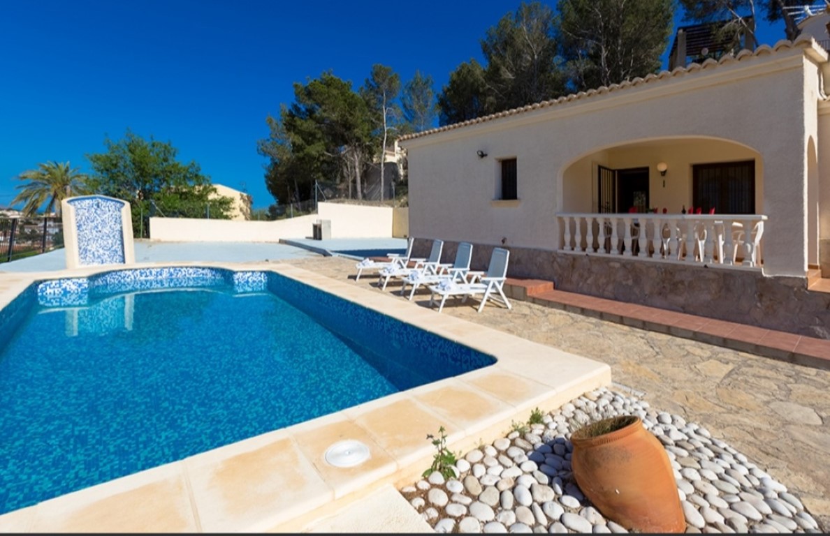 Exterior view featuring a blue mosaic pool, natural stone sun deck with loungers, and a traditional arched terrace with white balustrades.