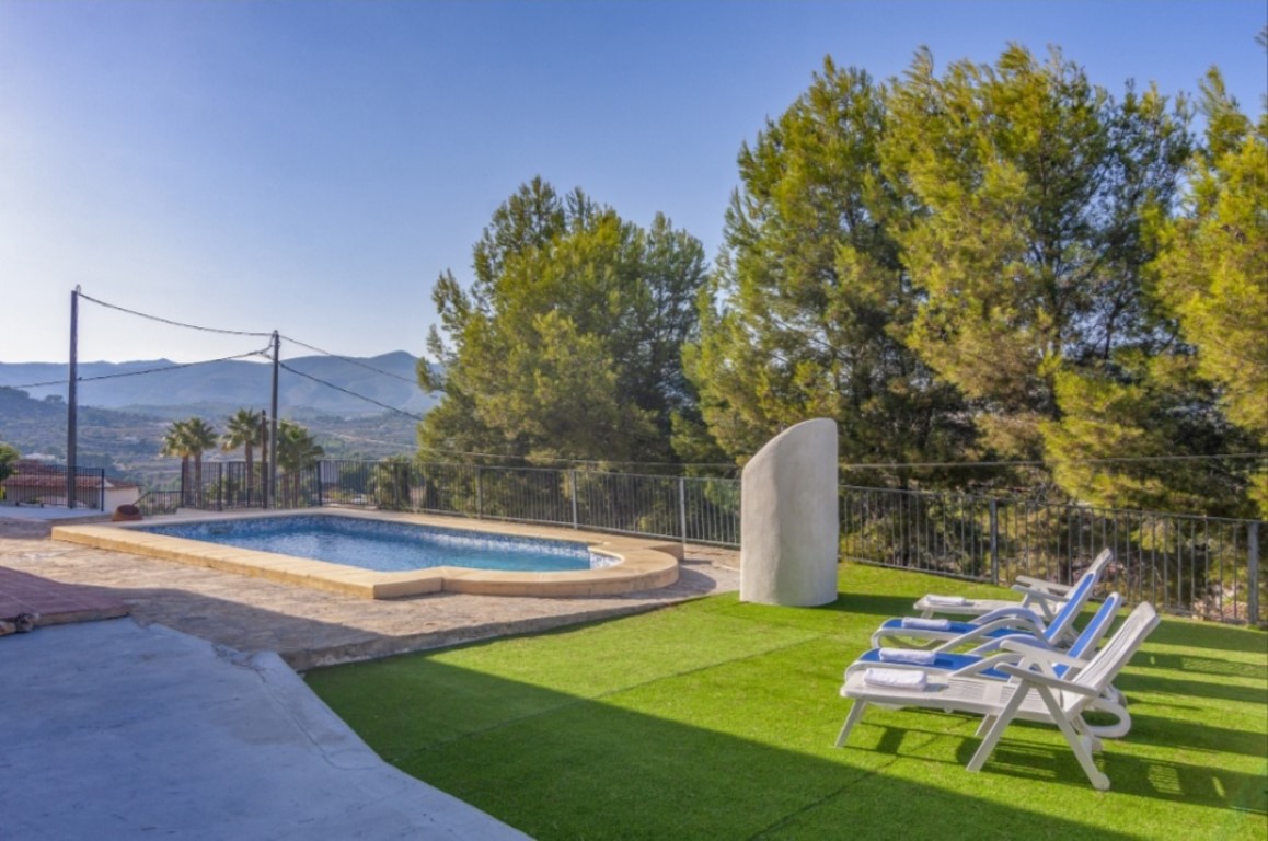 Outdoor pool area featuring a stone-paved terrace, artificial turf, and sun loungers, framed by mature pine trees and panoramic mountain views.