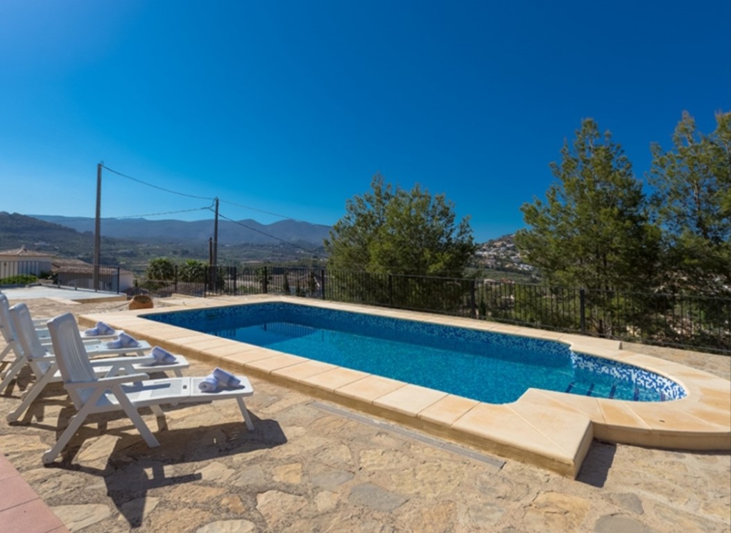 Rectangular swimming pool with blue mosaic tiling and side steps, bordered by a natural stone terrace with sun loungers and open mountain views.