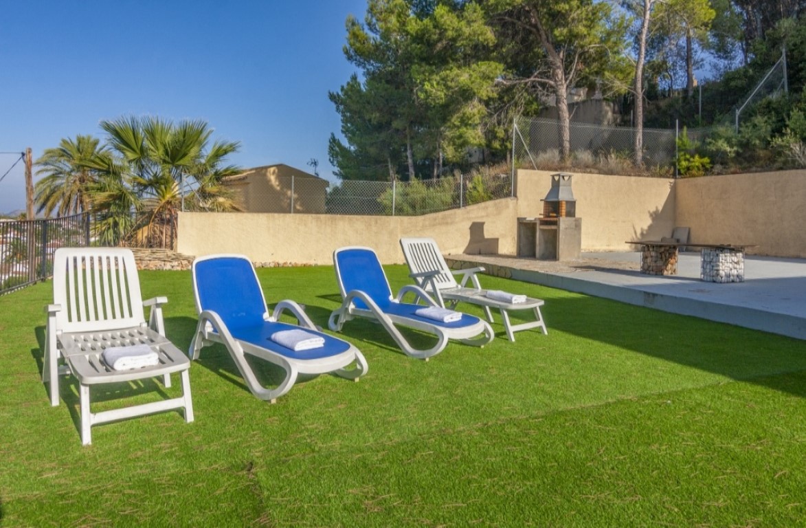 Terrace area with artificial lawn featuring four sun loungers, a built-in stone barbecue, and a gabion-style bench, surrounded by mature pine trees.