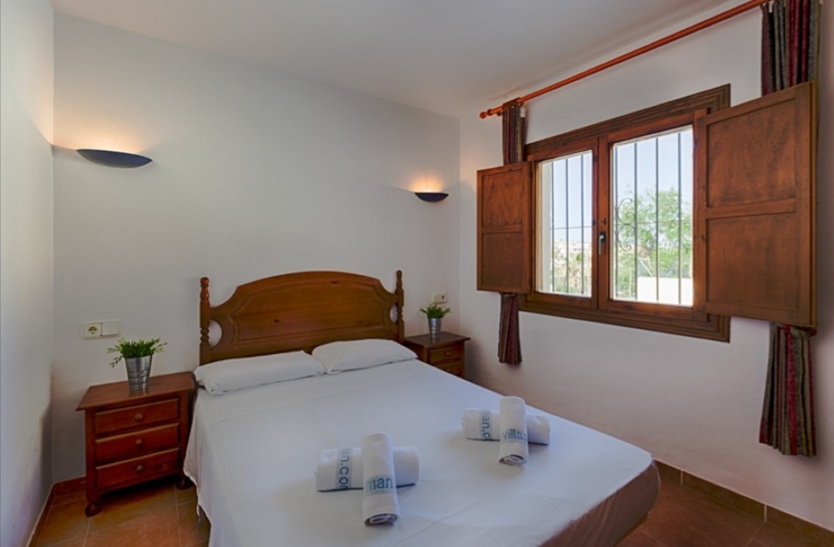 Traditional double bedroom featuring a wooden headboard, matching nightstands, wall sconces, and a window with classic wooden shutters and security bars.