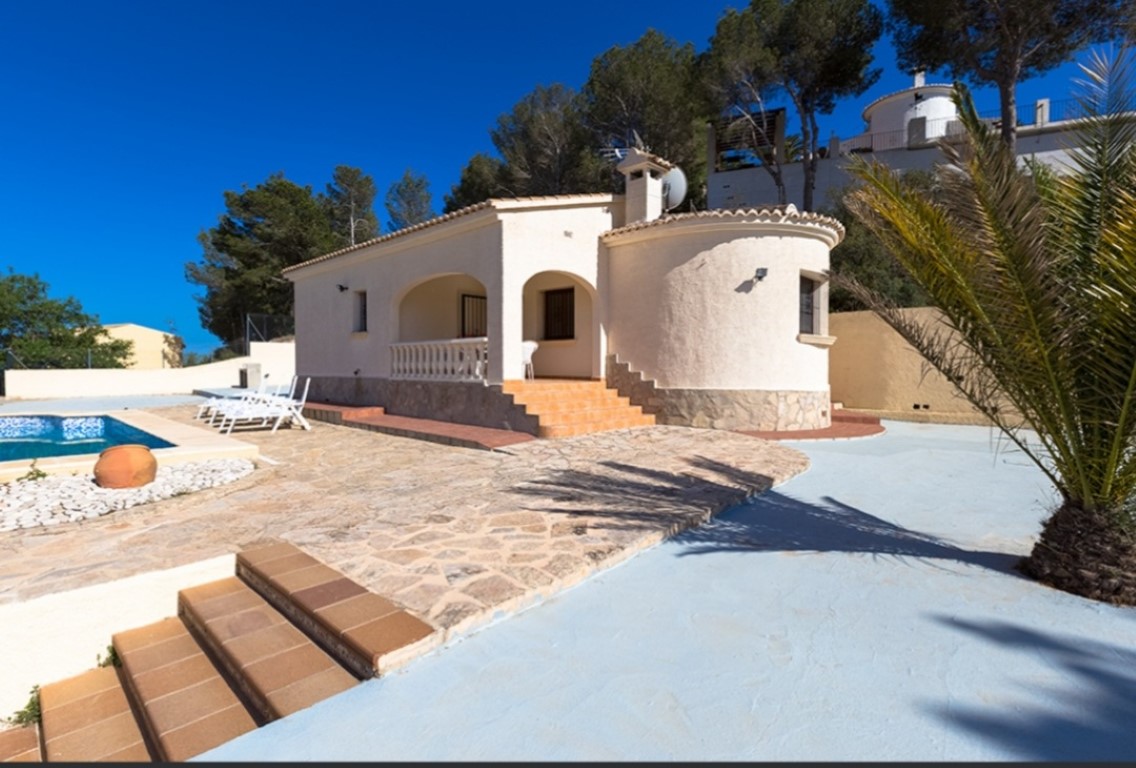 Traditional villa exterior featuring an arched porch, natural stone terrace, and private pool area set against a backdrop of mature pine trees.