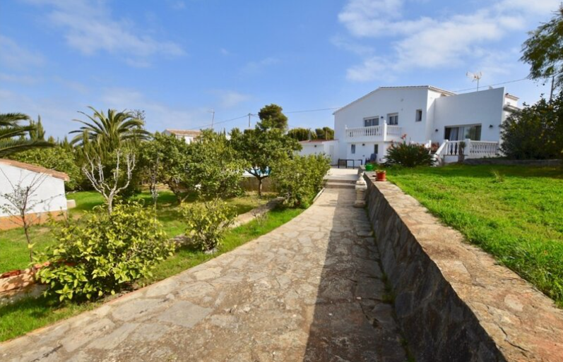A natural stone path leads through the Mediterranean garden towards the white villa, featuring manicured lawns, palm trees, and traditional balustrades.