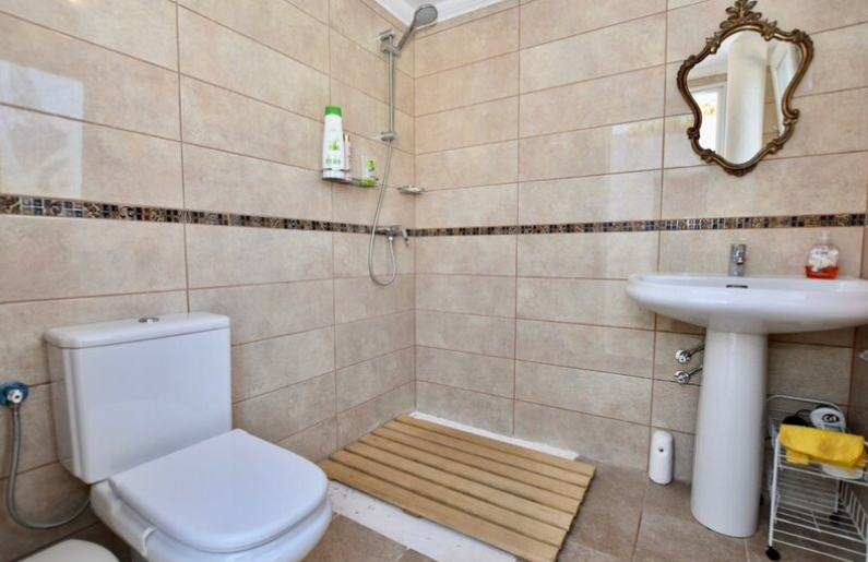Bathroom featuring a walk-in shower with a wooden slat floor mat, beige tiling with a mosaic border, and an ornate gold-framed mirror.