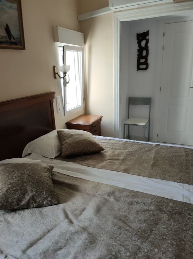 Bedroom featuring a double bed with a classic wooden headboard and air conditioning. It includes a bedside table, natural light from a side window, and a functional corner chair.