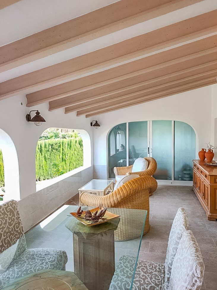 Covered naya featuring exposed ceiling beams, a glass-top dining set, and wicker lounge seating. Arched openings provide natural light and views of the Mediterranean greenery.
