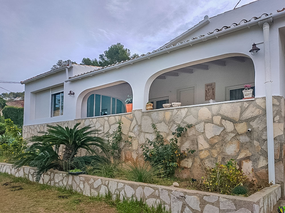 Exterior view of the villa featuring a traditional arched naya and natural stone masonry. The garden area includes Mediterranean plants and a white facade.