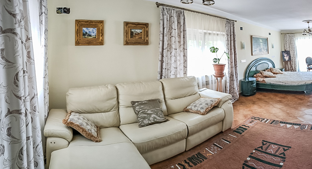 Guest house living area featuring a cream leather sofa, ceramic tile flooring, and an integrated bedroom space with classic decor and natural light.