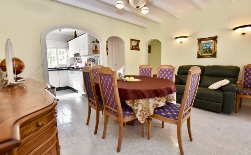 Guest house living-dining area featuring exposed ceiling beams, classic furniture, and an arched entrance to the white kitchen.