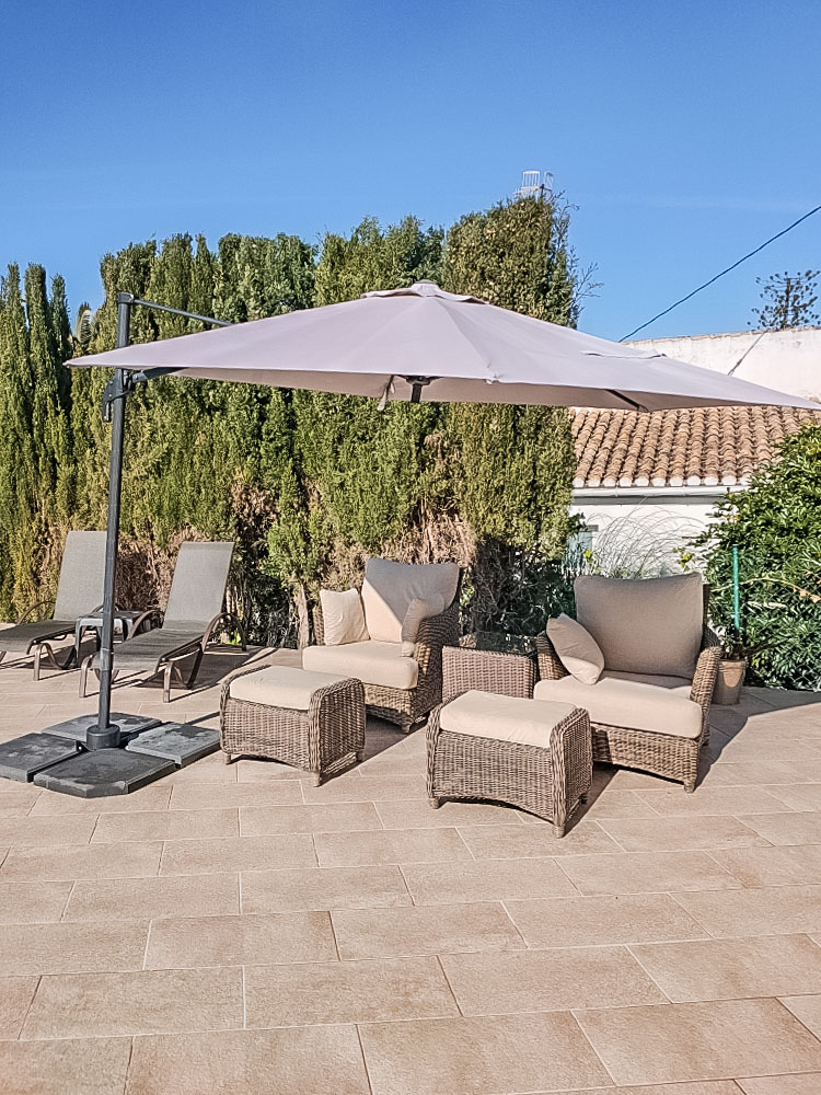 Outdoor lounge area featuring grey wicker armchairs with footstools and a large cantilever parasol on a stone-tiled terrace bordered by mature hedges.