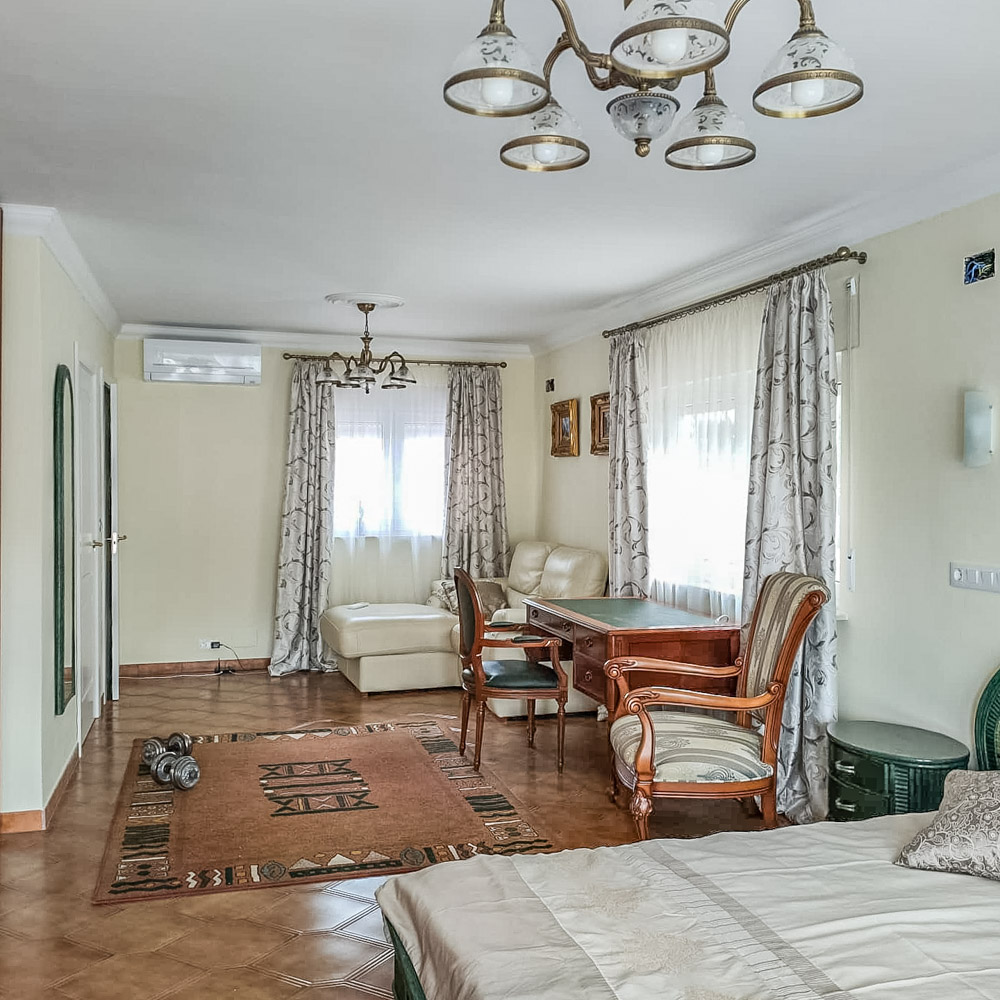 Spacious bedroom featuring a classic wooden desk, cream leather seating area, and air conditioning. Natural light fills the room through windows with floral drapery.
