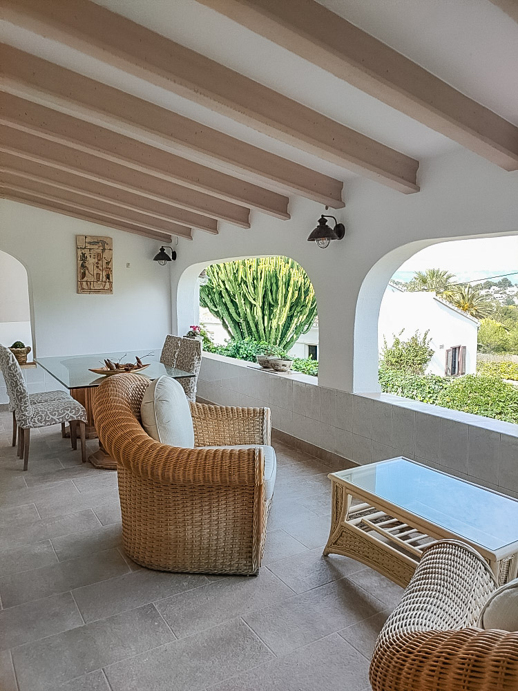 Spacious covered naya featuring exposed wooden beams, wicker lounge furniture, and traditional arches overlooking the Mediterranean garden.