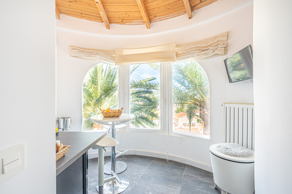 Breakfast nook featuring arched windows with palm tree views, a wooden beamed ceiling, and a high table with stools on dark tiled flooring.