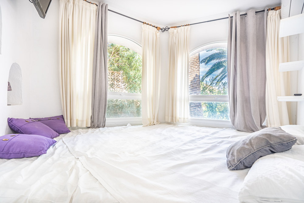 Bright bedroom featuring arched corner windows with views of palm trees and greenery. The space includes white linens, purple accents, and air conditioning.