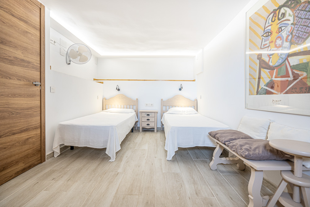 Bright bedroom featuring twin beds, wood-effect ceramic flooring, indirect ceiling LED lighting, and a functional wall-mounted fan.