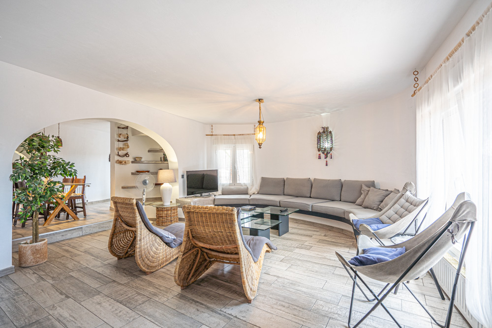 Bright living area featuring a built-in curved sofa, wicker lounge chairs, and wood-effect tiling. An architectural archway leads to the adjacent dining space.