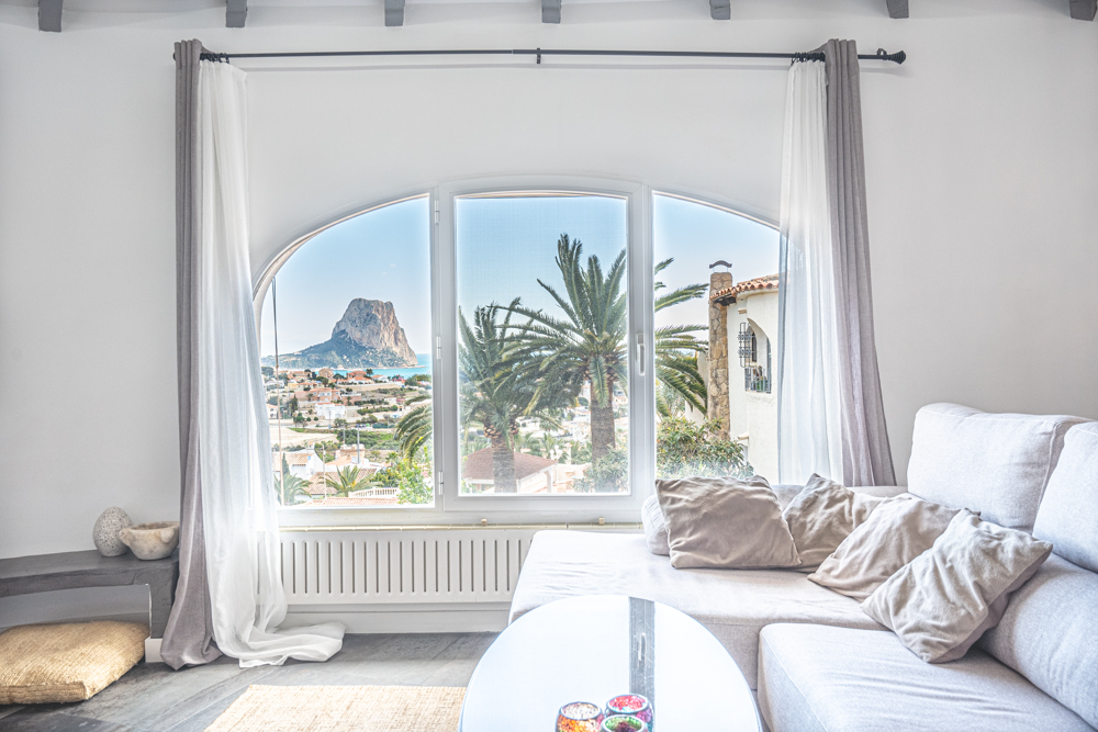 Bright living area featuring a large arched window with panoramic views of the Peñón de Ifach and the Mediterranean Sea, finished with exposed ceiling beams.