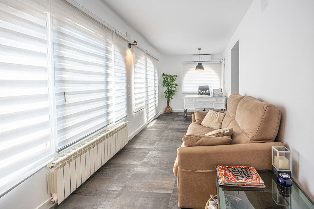 Bright naya featuring dark tile flooring, a beige sofa, and a dedicated workspace. Equipped with a radiator, air conditioning, and large windows with white zebra blinds.