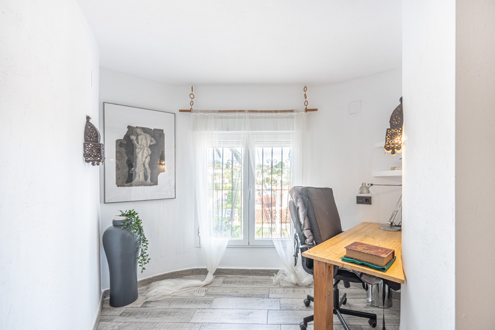 Bright study area featuring a wooden desk, wood-effect ceramic tiles, and a window with security bars providing natural light and ventilation.