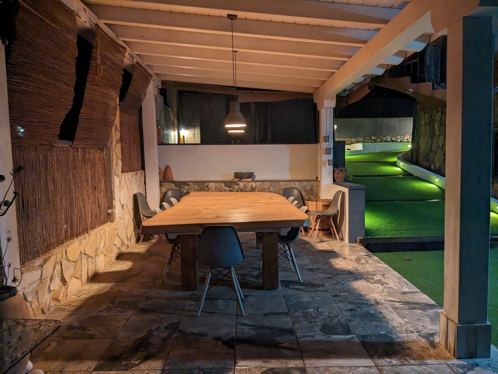 Covered outdoor dining area featuring a solid wood table, stone-clad walls, and a beamed ceiling, overlooking the illuminated garden path.
