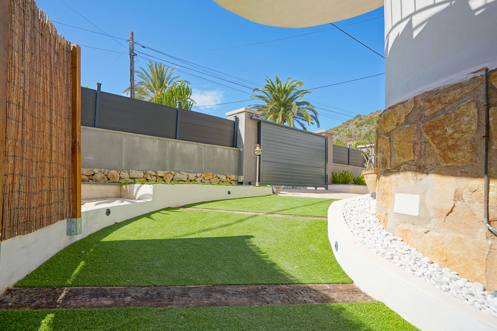 Exterior entrance featuring artificial turf with wooden sleepers and natural stone walls. Includes a modern sliding gate and decorative white gravel borders.