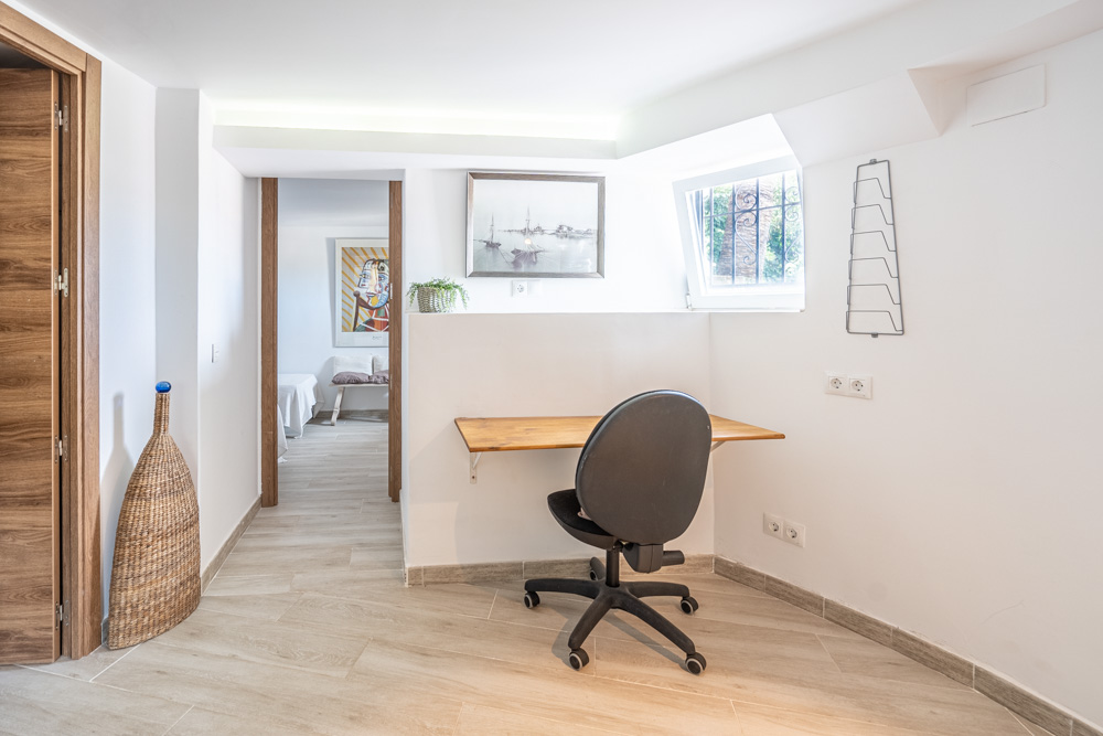 Integrated office nook featuring a wooden desk, ergonomic chair, and recessed LED lighting. The space includes wood-effect porcelain flooring and leads to the bedrooms.