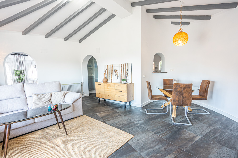 Living and dining space featuring exposed grey ceiling beams, dark tile flooring, and arched doorways. Includes a modern wooden sideboard and a glass-top dining table.