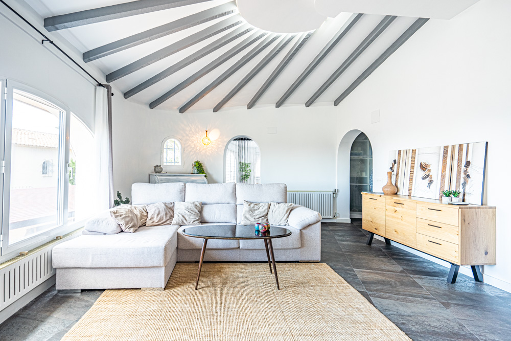 Living area featuring a radial grey beamed ceiling and curved walls. Includes a light grey sectional sofa, dark tile flooring, and distinctive arched doorways.