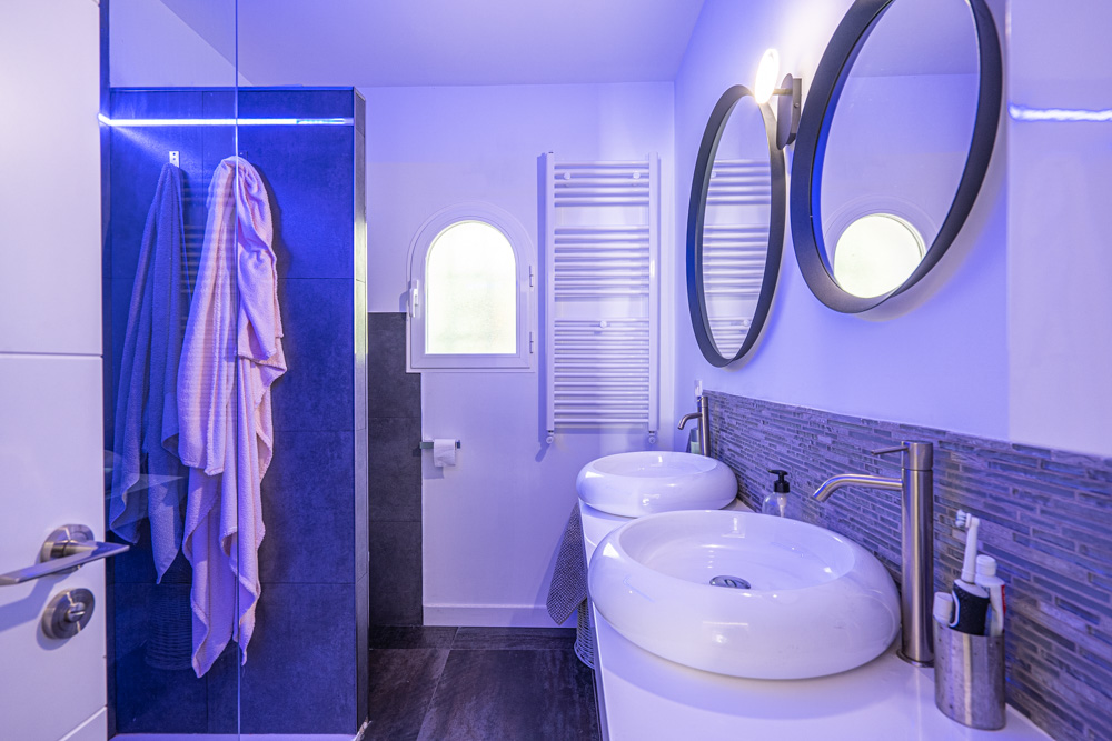 Modern bathroom featuring a double vanity with round vessel sinks, circular mirrors, and a walk-in shower with atmospheric blue LED lighting.