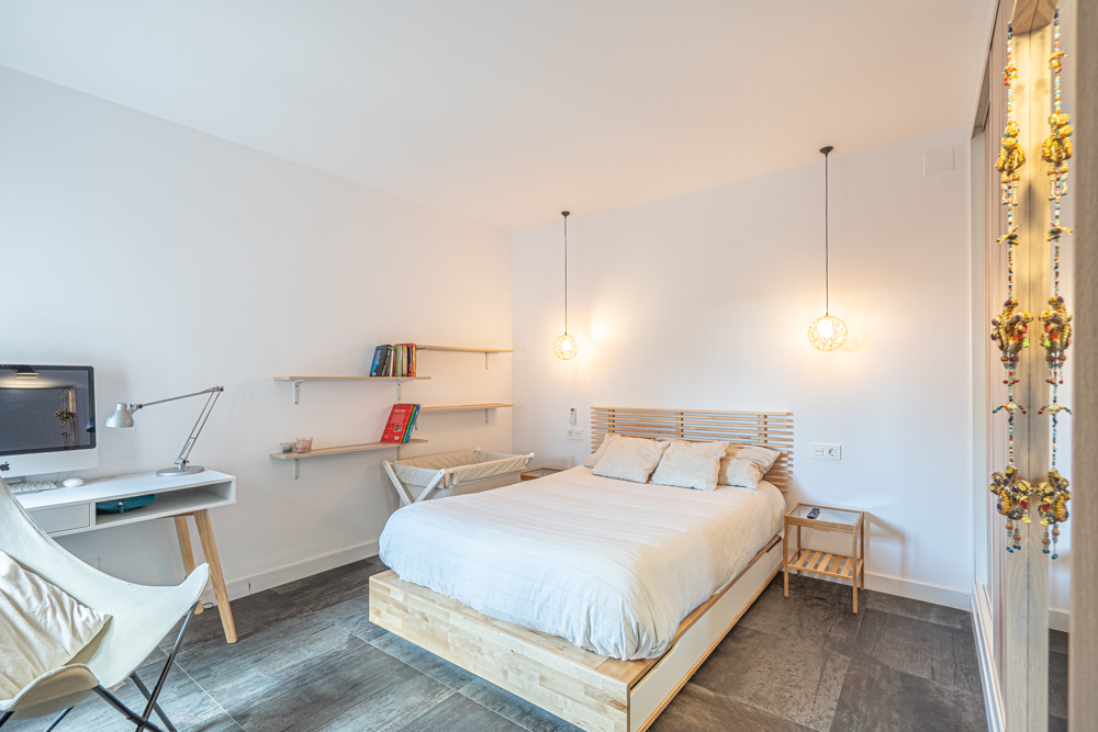Modern bedroom featuring a wooden bed frame, integrated workspace with a white desk, and floating shelves. Includes designer pendant lighting and dark tile flooring.