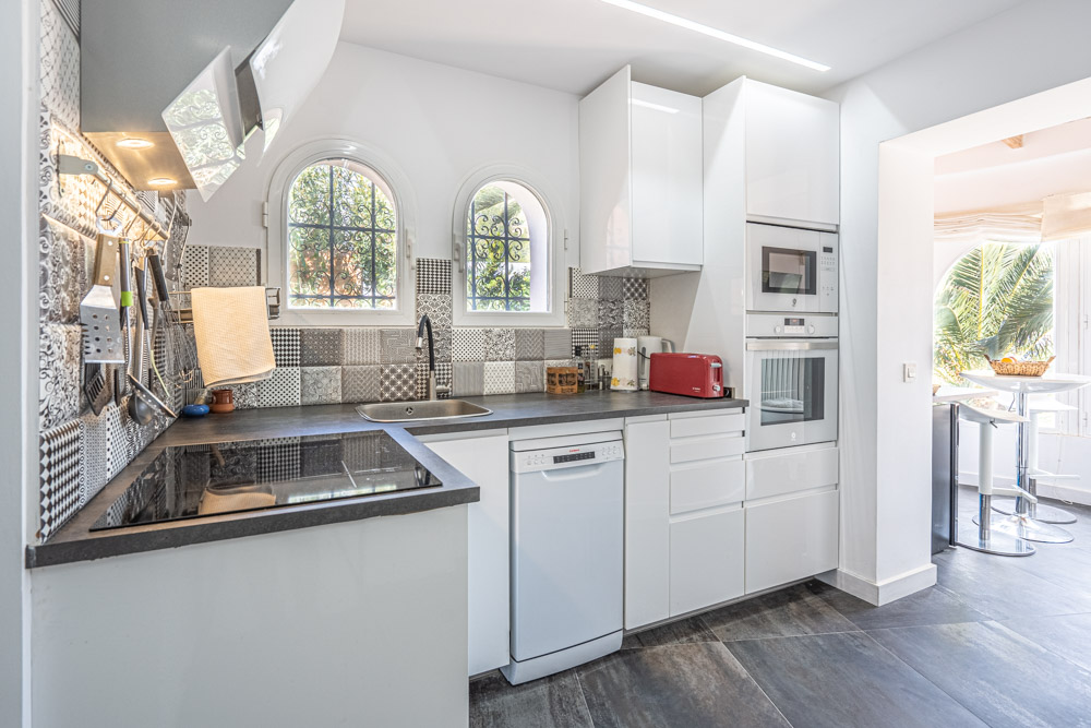 Modern kitchen featuring white gloss cabinetry, dark countertops, and a patterned tile backsplash. Equipped with integrated appliances and arched windows providing natural light.