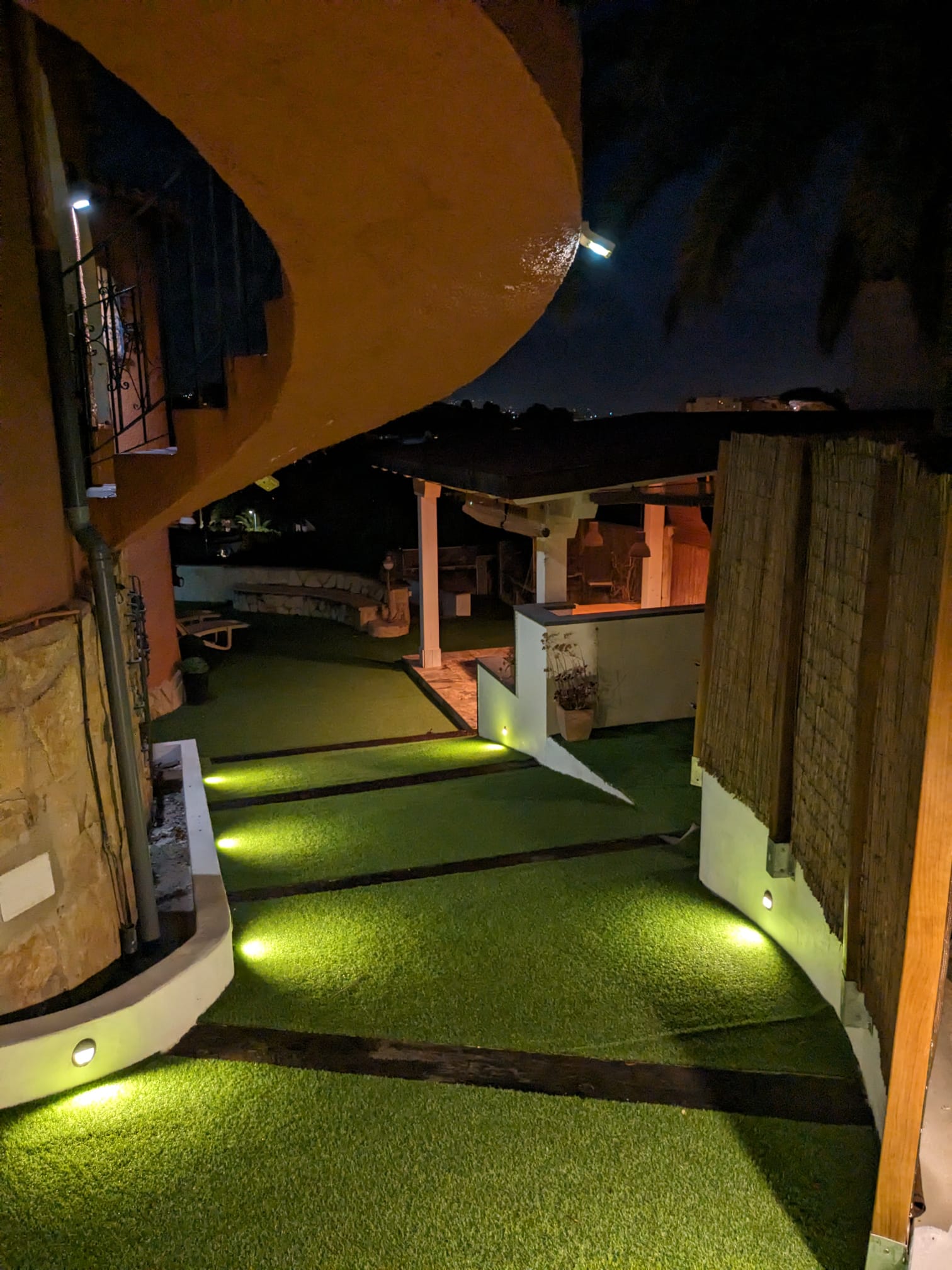 Night view of the exterior pathways featuring integrated LED floor lighting, artificial turf, and a covered pergola area in the background.