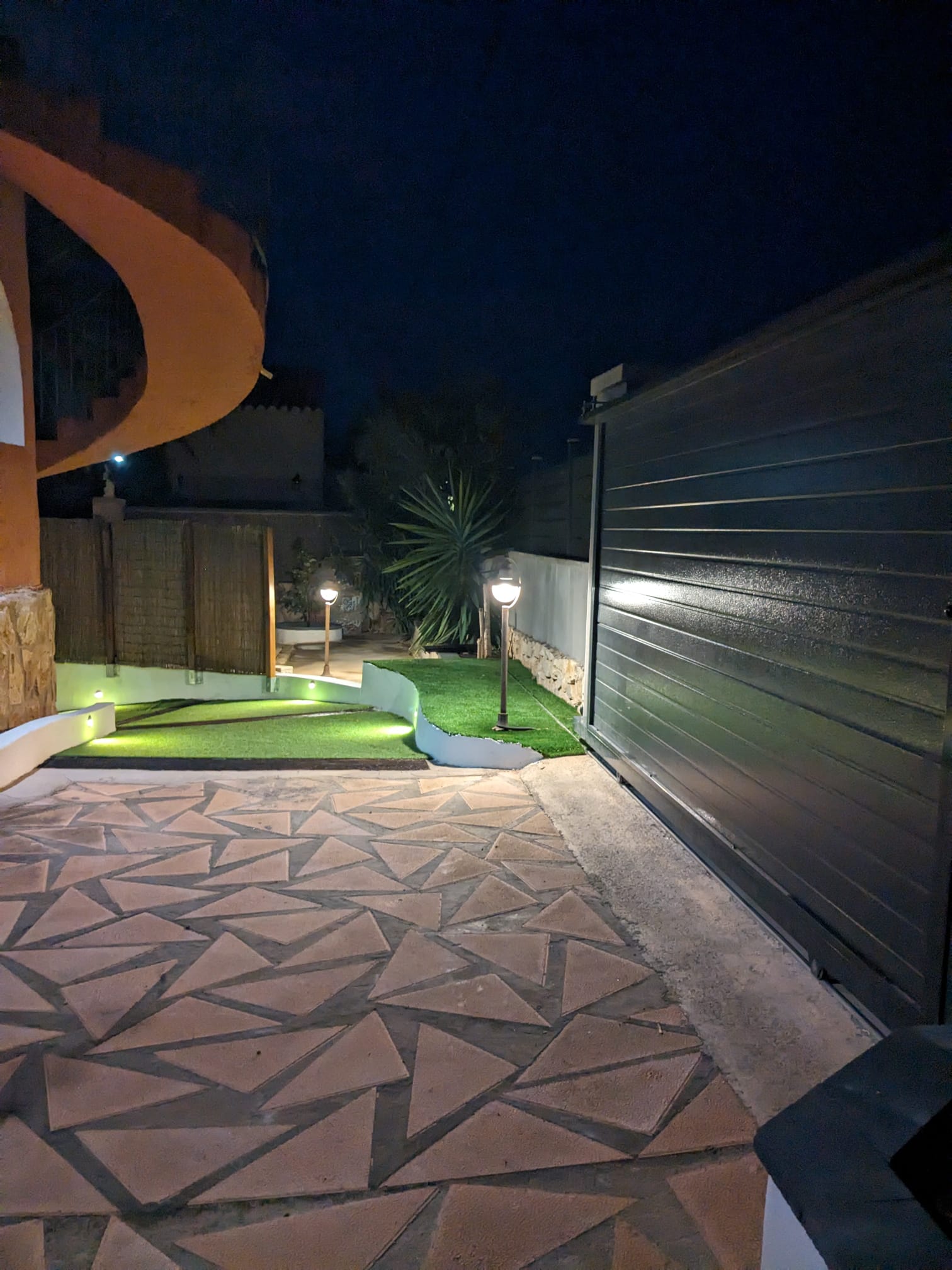 Nighttime exterior entrance featuring a grey sliding gate, geometric stone paving, and integrated lighting along the artificial turf and walls.