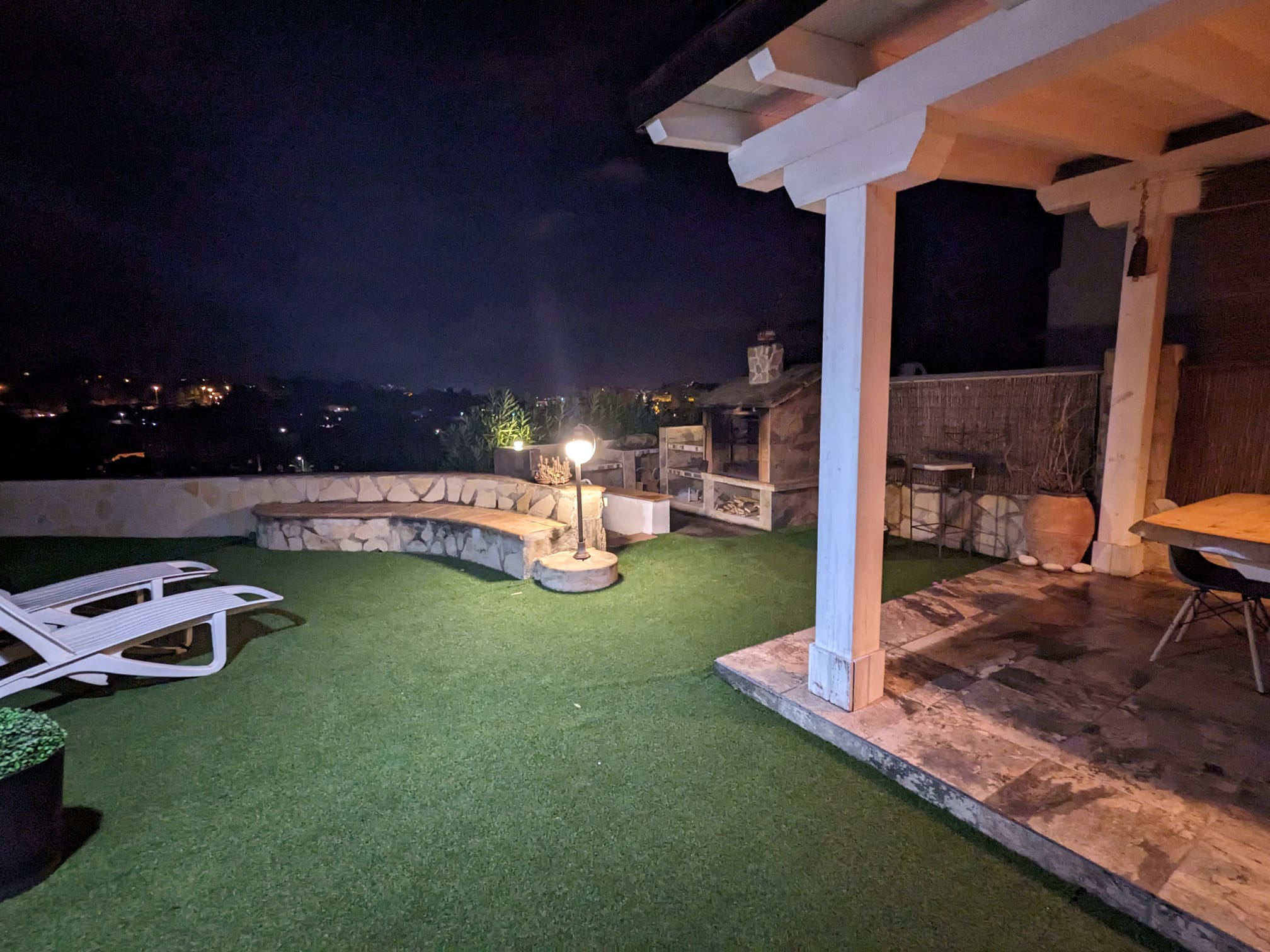 Nighttime outdoor area featuring a stone barbecue, curved masonry bench, and a wooden pergola with a dining set adjacent to artificial turf and loungers.
