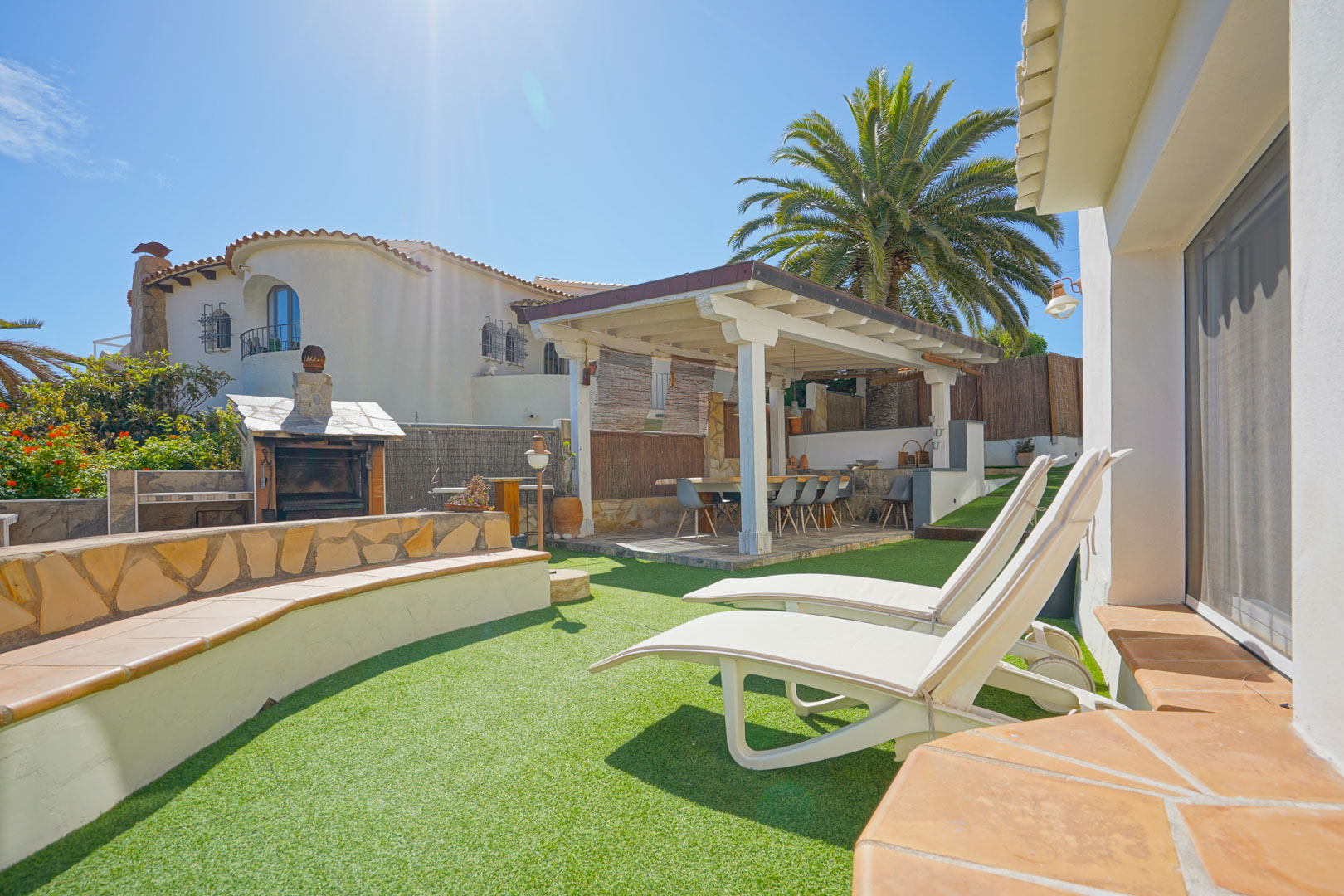 Outdoor area featuring artificial grass, white sun loungers, and a wooden pergola covering an alfresco dining space next to a stone barbecue.