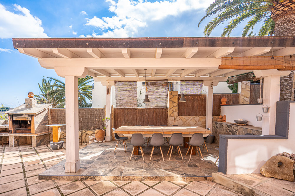Outdoor dining area featuring a white wooden pergola, a large solid wood table with modern seating, and a stone-built barbecue station on a tiled terrace.