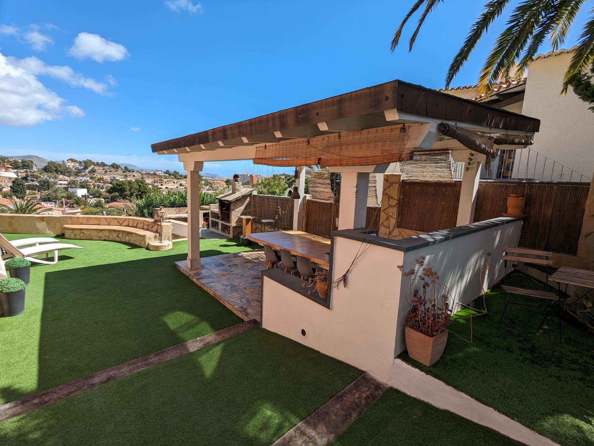Outdoor dining area featuring a wooden pergola, large table, and integrated stone barbecue, set against an artificial lawn with views of the Calpe landscape.