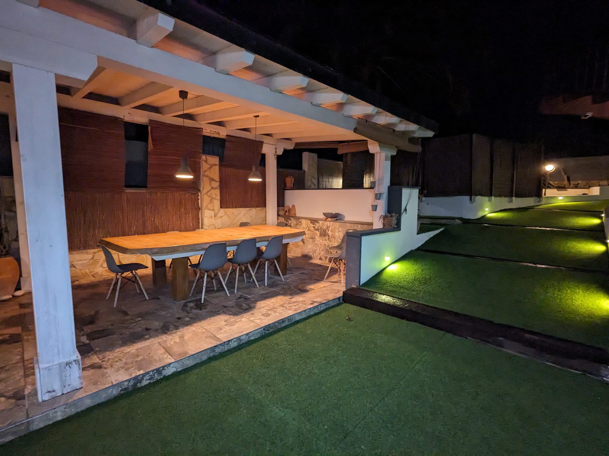 Outdoor dining area under a white wooden pergola featuring a large timber table, modern seating, and stone flooring adjacent to an illuminated lawn.